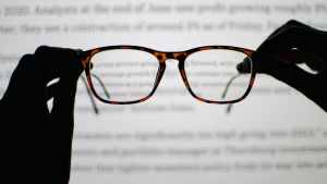 POV picture of hands holding up glasses for inspection in front of a computer monitor. The computer is out of focus, and the hands and glasses appear as a silhouette due to the brightness of the computer monitor.